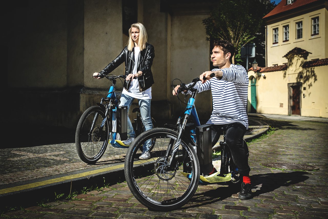 Two people on an electric scooter, gazing toward the horizon, riding along the cobblestone streets of Prague