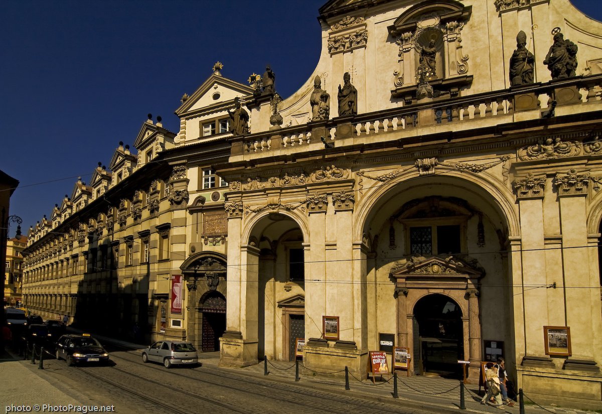 The carved stone facade of a large Baroque monument, flanked by a cobblestone street with a few cars passing by.