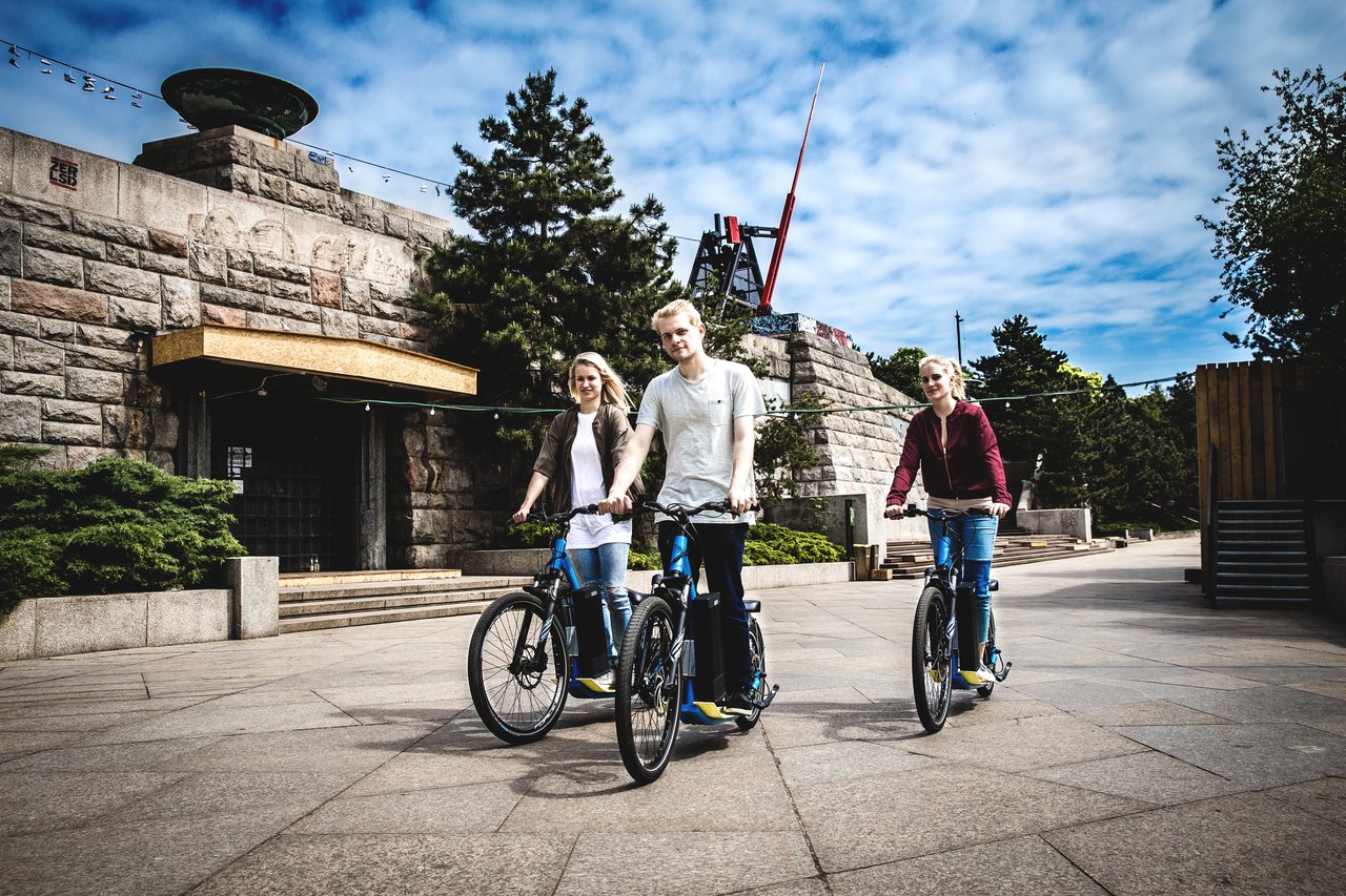 Three people on electric scooters in front of a stone building topped by a metronome